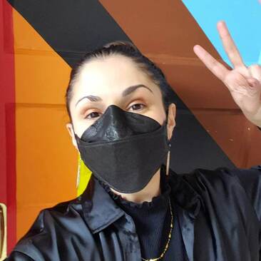 Leah wears a black N95 mask and poses in front of the door to Calgary's Outlink Centre, which is painted in the colours of the Progress Pride flag. She holds up a peace sign, and wears her dark brown hair in a bun. You can tell she's smiling behind her mask! 